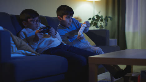 Two-Young-Boys-Sitting-On-Sofa-At-Home-Playing-With-Computer-Games-Console-On-TV-Holding-Controllers-Late-At-Night-3